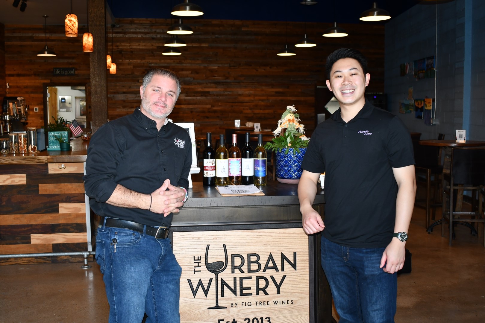 Two men in Urban Winery shirts stand beside the reception area podium. The wine bar and seating is visible in the background.